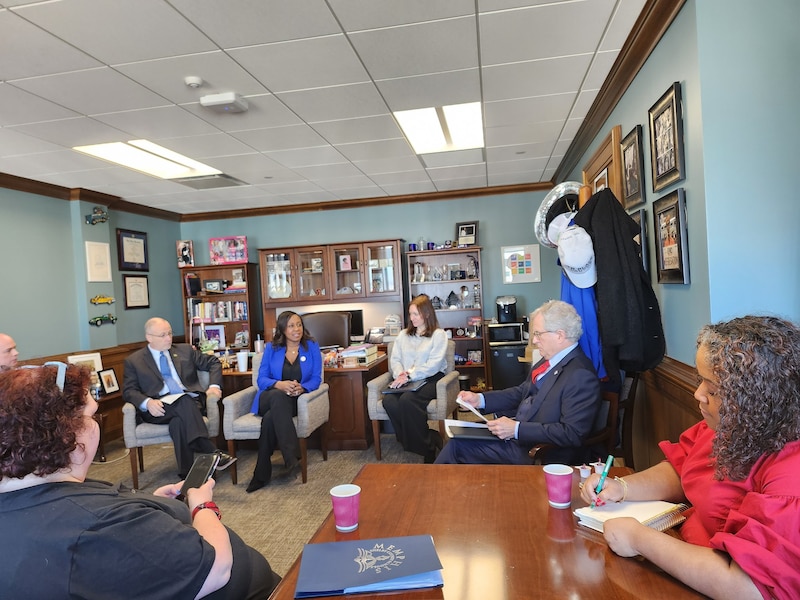 A group of people wearing business clothes sit in chairs in an office with blue walls.