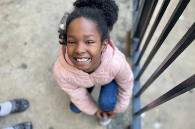 A young girl with dark hair and wearing a pink coat bends over to tie her shoe and looks up at the camera smiling.