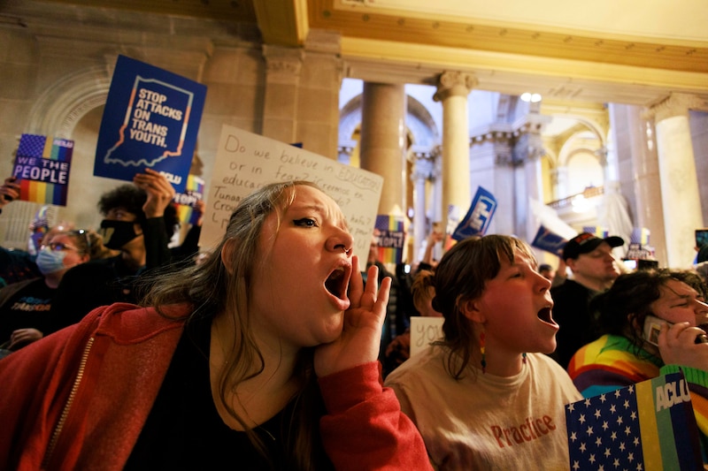 Two protestors chant from a crowd inside a large stone building with signs and people in the background.