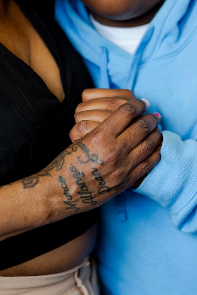 A Black mother's hand embraces her son's hand. Her hand has some tattoos and painted, long nails. A blue sweater in the background.