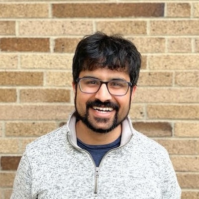 Headshot of a man with black hair and a black beard wears a gray sweater