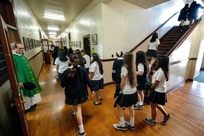 Teenage students in plaid skirts and white shirts walk through a school hallway and up a staircase. A clergyman in a green robe watches from a doorway.