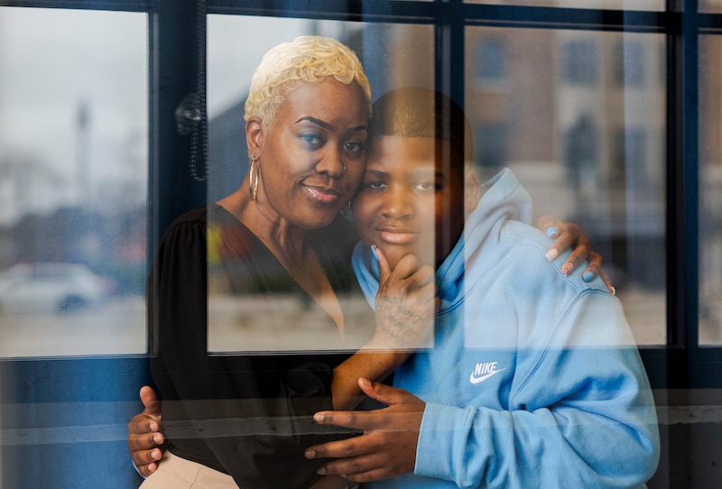 A Black mother and son pose for a portrait standing next to each other embracing against a blue curtain.