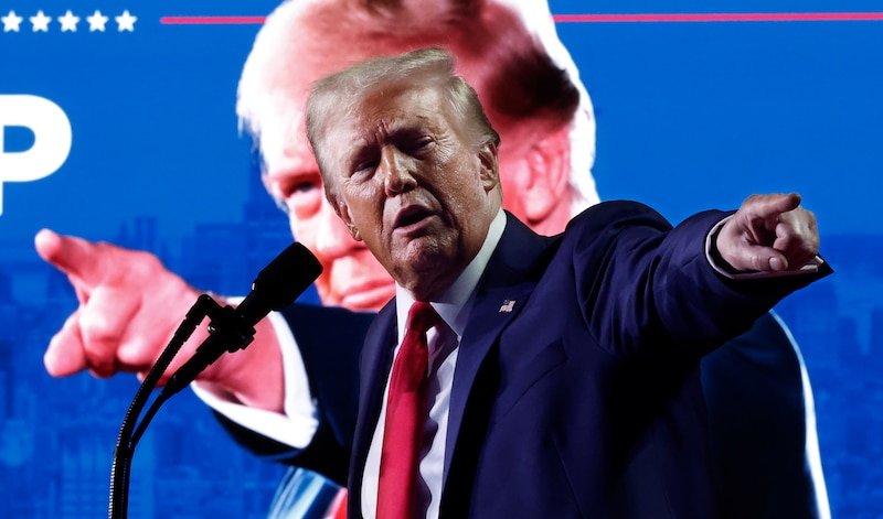 A man in a blue suit and a red tie stands pointing behind a podium and in front of a photo of himself on a projector in the background.