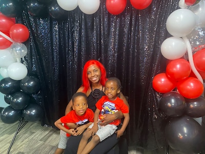 A woman holds two young boys in front of a black background with red, white and black balloons in an arch.